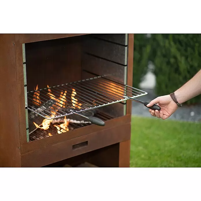 Westmann Grillkamin - L x B x H: 410 x 700 x 2.290 mm, Rost, Stahl - Eine Hand platziert einen silbernen Grillrost in einen beleuchteten, rostfarbenen Außenkamin, bereit zum Grillen.