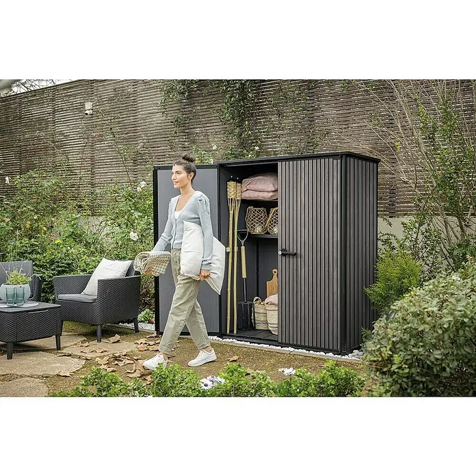 Keter Geräteschrank Signature Venetian High Store - Außenmaß inkl. Dachüberstand (B x T): 140 x 73,6 cm, Kunststoff, Grau - Dunkelgrauer Gartenschrank im Garten mit einer Frau, die Kissen und eine Decke trägt, Gartenmöbeln und viel Grün.