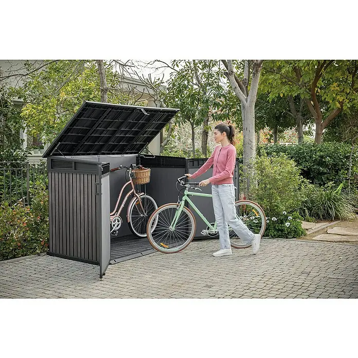 Keter Truhe Signature Venetian Grande StoreL x B x H: 109,3 x 190,5 x 132 cm, Grau - Frau schiebt Fahrrad in eine braune Fahrradaufbewahrungsbox mit offenem Deckel. Praktische Aufbewahrungslösung für Fahrräder im Außenbereich.