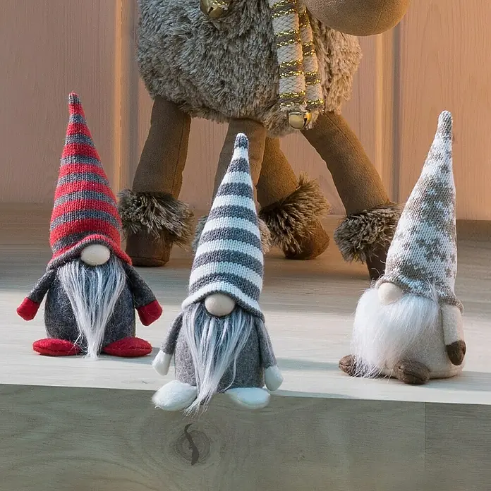 Dekofigur Mini Wichtel - L x B x H: 7,7 x 6,7 x 18 cm, Textil, Weihnachtswichtel - Dekoratives Bild von drei Stoffgnomen mit gestreiften Mützen und weißen Bärten vor einem weichen Rentier.