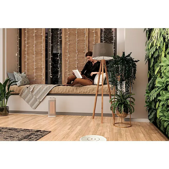 Voltomat HEATING Keramik-Heizlüfter Mini Tower - 2.000 W, Silber, 20,5 x 16,2 x 44,5 cm - Frau liest auf einer Fensterbank, eine Stehlampe und eine grüne Wandbelebung im Raum, während ein Staubsaugerroboter den Boden reinigt.