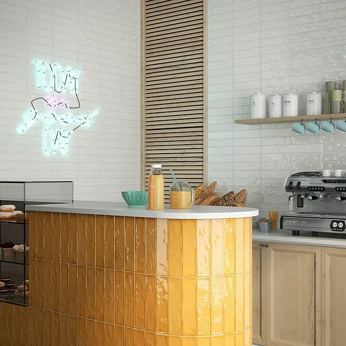 BHS Showroom Revestimiento de pared Rain - 7,5 x 30 cm, Bianco, Brillante - Un mostrador con azulejos verticales naranjas que crea un ambiente cálido y acogedor en una cafetería o panadería.