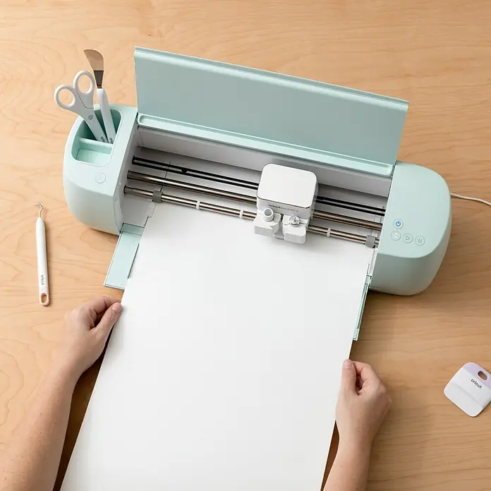 Cricut Aufkleber Smart Label Auflösbar1 Stk., Weiß, 33 x 61 cm - Cricut Schneideplotter in Mintgrün mit Werkzeugen und einer Person, die ein Blatt Papier einlegt, für präzise Bastelarbeiten.