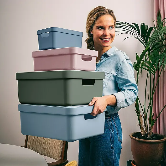 Sunware Aufbewahrungsbox Sigma Home - L x B x H: 35 x 24,6 x 12,2 cm, Kunststoff, Rosa - Frau trägt einen Stapel bunter Aufbewahrungsboxen. Die Boxen sind in Blau-, Rosa- und Grüntönen gehalten und haben praktische Griffe.