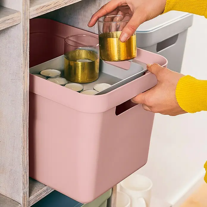 Sunware Aufbewahrungsbox Sigma Home - L x B x H: 35 x 24,6 x 24,3 cm, Kunststoff, Rosa - Nahaufnahme einer Person, die einen rosa Aufbewahrungskorb mit Teelichtern und goldenen Gläsern hält. Praktische Aufbewahrungslösung.