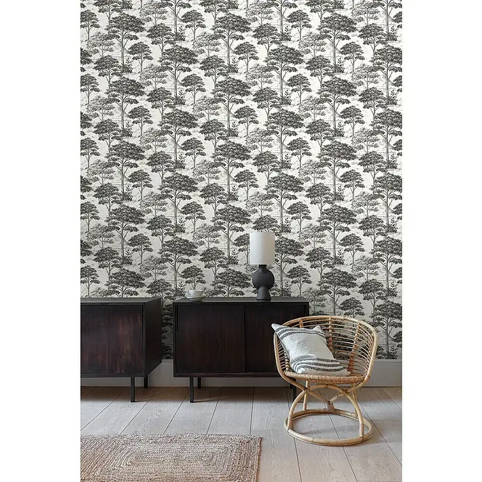 Papel pintado Arboles B&N - Blanco/Negro, 10 x 0,53 m - Primer plano de dos armarios de madera oscura y una silla de mimbre con cojín frente a un papel tapiz con motivos de bosque.