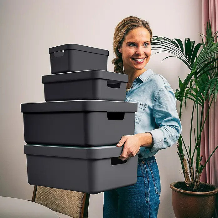 Sunware Aufbewahrungsbox Sigma Home - L x B x H: 44,5 x 34,5 x 18,3 cm, Kunststoff, Anthrazit - Frau trägt einen Stapel grauer Aufbewahrungsboxen mit weißen Deckeln. Ideal für Organisation und Lagerung im Haushalt.