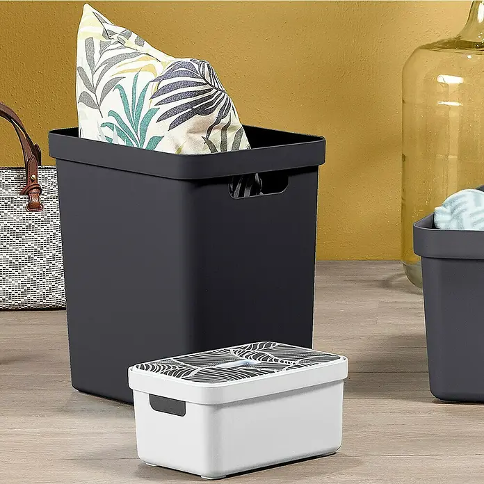 Sunware Aufbewahrungsbox Sigma Home - L x B x H: 35 x 24,6 x 36,3 cm, Kunststoff, Schwarz - Ein grauer Aufbewahrungskorb mit Kissen und eine weiße Box mit Muster stehen auf einem Holzboden vor einer gelben Wand.