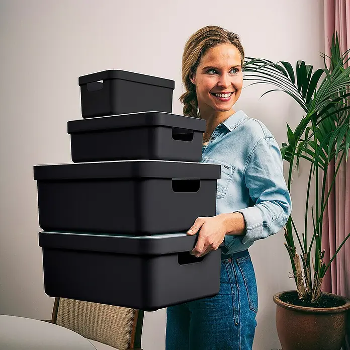 Sunware Aufbewahrungsbox Sigma Home - L x B x H: 44,5 x 34,5 x 18,3 cm, Kunststoff, Schwarz - Frau trägt einen Stapel schwarzer Aufbewahrungsboxen mit weißen Deckeln.
