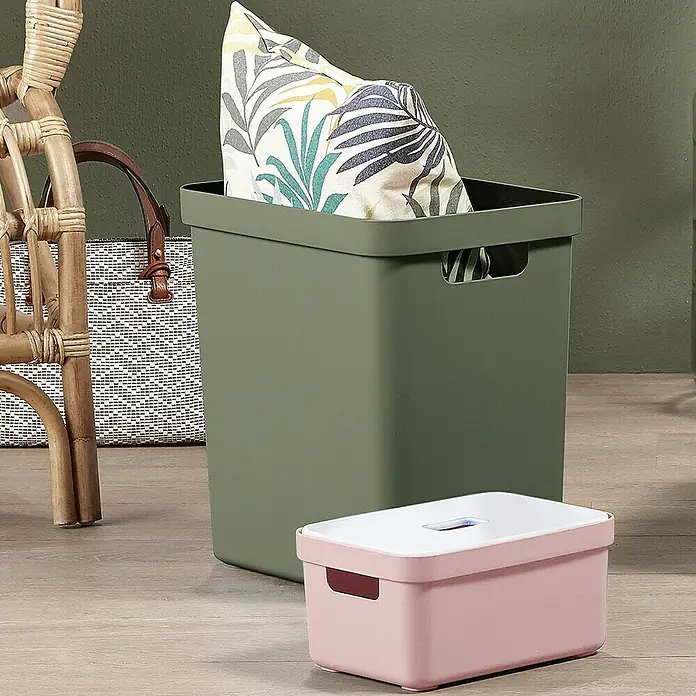 Sunware Aufbewahrungsbox Sigma Home - L x B x H: 35 x 24,6 x 36,3 cm, Kunststoff, Dunkelgrün - Einrichtungsgegenstände: Ein olivgrüner Aufbewahrungskorb mit einem Kissen darin und ein rosa Aufbewahrungsbehälter mit weißem Deckel stehen auf dem Boden.