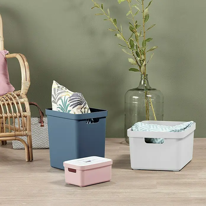 Sunware Aufbewahrungsbox Sigma Home - L x B x H: 44,5 x 34,5 x 24,3 cm, Kunststoff, Weiß - Eine Nahaufnahme von drei dekorativen Aufbewahrungsboxen in Blau, Rosa und Weiß auf einem Holzboden. Im Hintergrund sind Pflanzen und ein Rattansessel zu sehen.