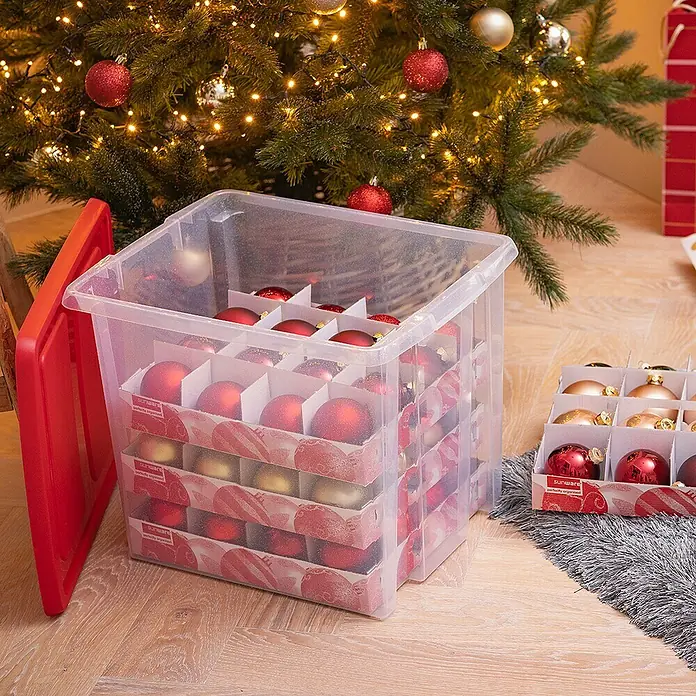 Sunware Aufbewahrungsbox Nesta für Weihnachtskugeln - Passend für: 48 Christbaumkugeln ( Ø 70 - 80 mm), L x B x H: 48 x 36,5 x 36,5 cm, Kunststoff, Transparent - Aufbewahrungsbox mit rotem Deckel und Fächern, gefüllt mit roten und goldenen Weihnachtskugeln, vor einem geschmückten Weihnachtsbaum.