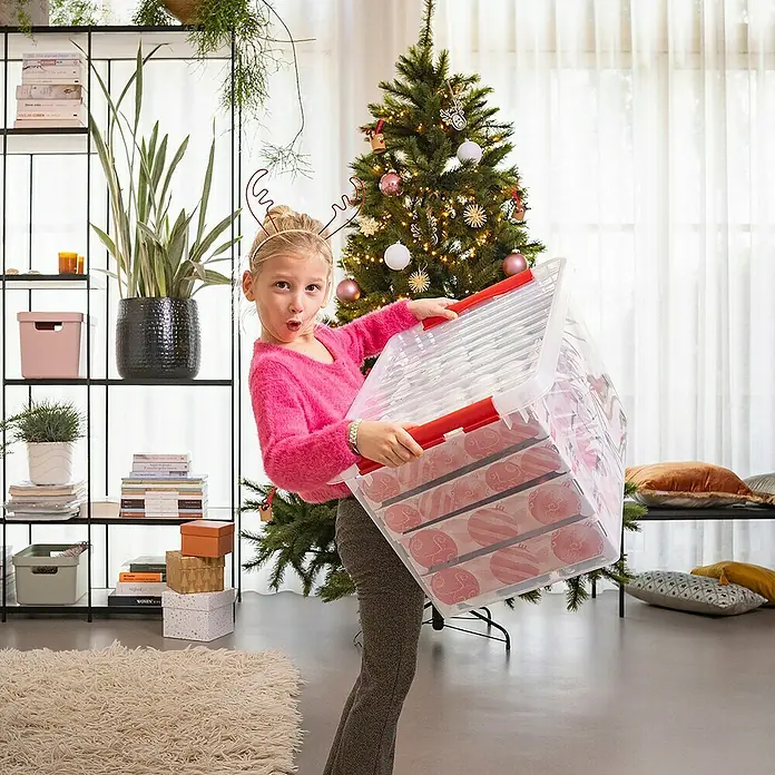 Sunware Aufbewahrungsbox Q-Line für Weihnachtskugeln - Passend für: 116 Christbaumkugeln (80 x Ø 45 - 60 mm & 36 x Ø 70 - 80 mm), L x B x H: 60 x 40 x 34 cm, Kunststoff, Transparent - Ein Mädchen trägt eine transparente Aufbewahrungsbox mit roten Griffen, gefüllt mit Weihnachtskugeln, vor einem Weihnachtsbaum.