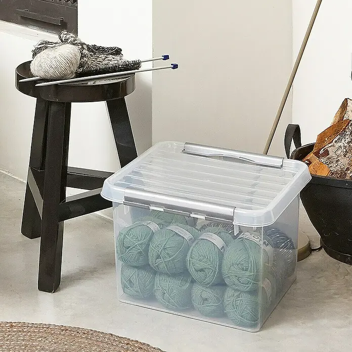 Sunware Aufbewahrungsbox Q-Line - L x B x H: 40 x 40 x 26 cm, Kunststoff, Transparent, Farbe Griff: Schwarz - Ein durchsichtiger Kunststoffbehälter gefüllt mit grüner Wolle steht auf dem Boden, daneben ein Hocker mit Strickzeug.