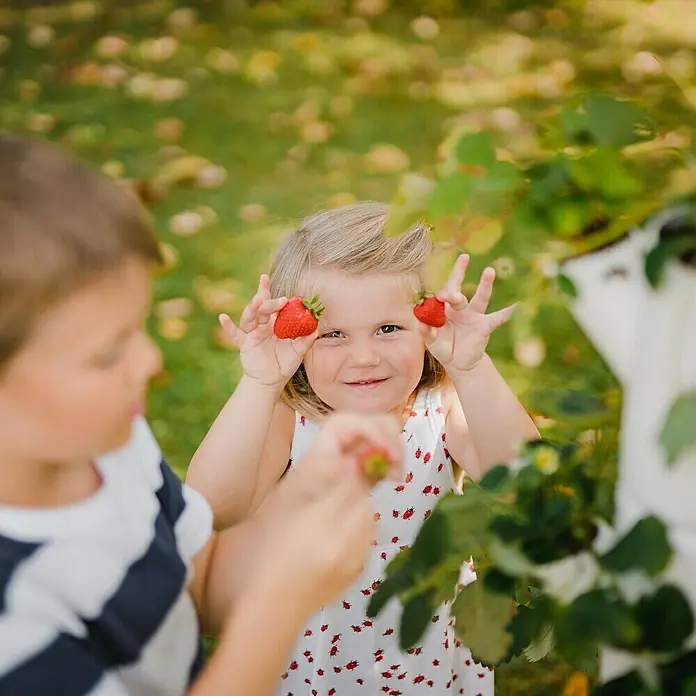 Gusta Garden Pflanzturm Sissi Strawberry Classic - 270 x 270 x 1.130 mm, Weiß - Dieses Bild zeigt zwei Kinder, die Erdbeeren pflücken. Das Mädchen hält zwei Erdbeeren vor ihr Gesicht und lächelt.