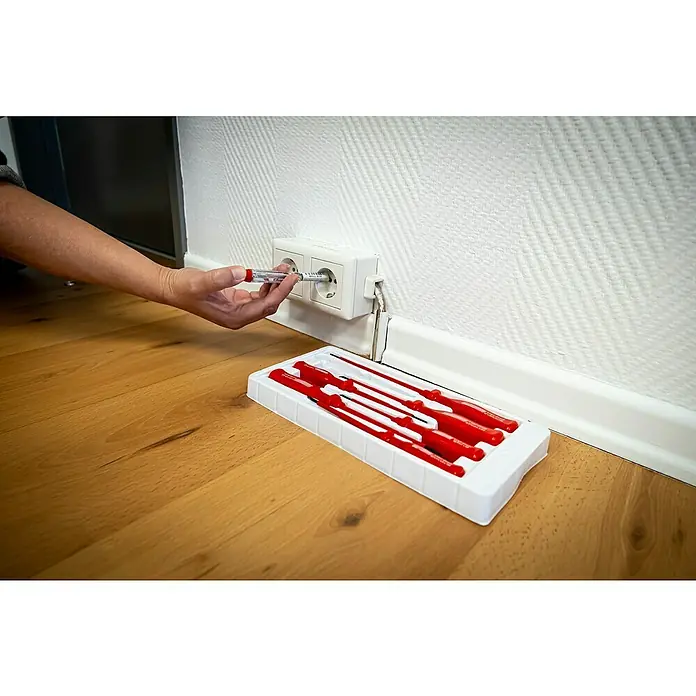 Brüder Mannesmann Elektriker-Schraubendreher - 7 -tlg. - Eine Hand, die einen Schraubendreher in eine Steckdose steckt, neben einem Set roter Schraubendreher auf einem Holzboden, vor einer weißen Wand.