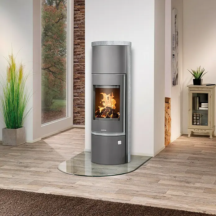Justus Kaminofen Faro W+ 2.0 - 7 kW, Raumheizvermögen: 144 m³, Verkleidung: Gusseisen/Speckstein, Gussgrau - Nahaufnahme eines Justus Kaminofens mit Glasbodenplatte, der Wärme und Eleganz in den Raum bringt. Das lodernde Feuer sorgt für eine gemütliche Atmosphäre.