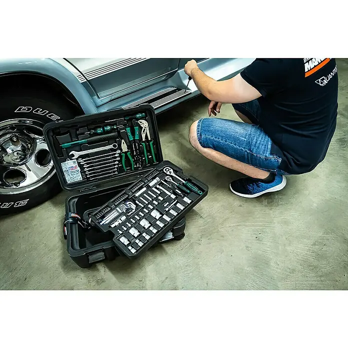 Brüder Mannesmann Werkzeugkoffer Green Line - L x B x H: 27 x 39 x 57 cm, 122 -tlg. - Werkzeugkasten neben einem Auto bei der Reparatur. Der Kasten ist voll mit Werkzeugen, darunter Schraubenschlüssel, Zangen und Schraubendreher.