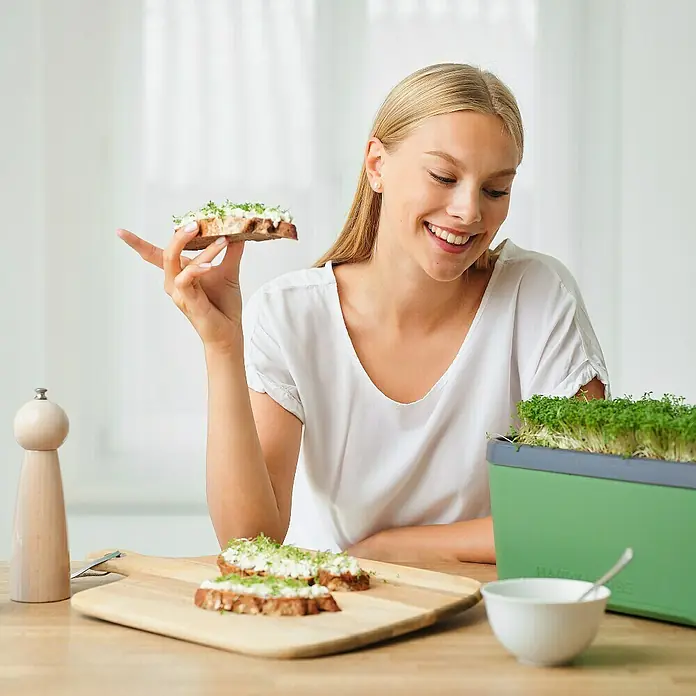 Gusta Garden Kräutertopf Harry Herbs - Kunststoff, Dunkelgrün - Eine lächelnde Frau genießt ein mit Kresse belegtes Brot, die frische Kresse stammt aus einem grünen Kresse-Anzuchtset.