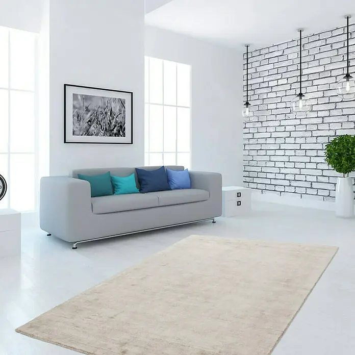 Kayoom Kurzflorteppich Luxury - Elfenbein/Taupe, 150 x 80 cm, 100 % Viskose - Beigefarbener Teppich in einem modernen Wohnzimmer mit grauer Couch, blauen Kissen und einer weißen Ziegelwand mit Pendelleuchten.