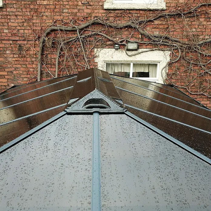 Palram – Canopia Pérgola estándar con cortinas Martinique - An x Pr: 360 x 500 cm, Color del armazón: Antracita, Aluminio - Primer plano del techo de un pabellón cubierto de gotas de lluvia, con una pared de ladrillo y hiedra al fondo.