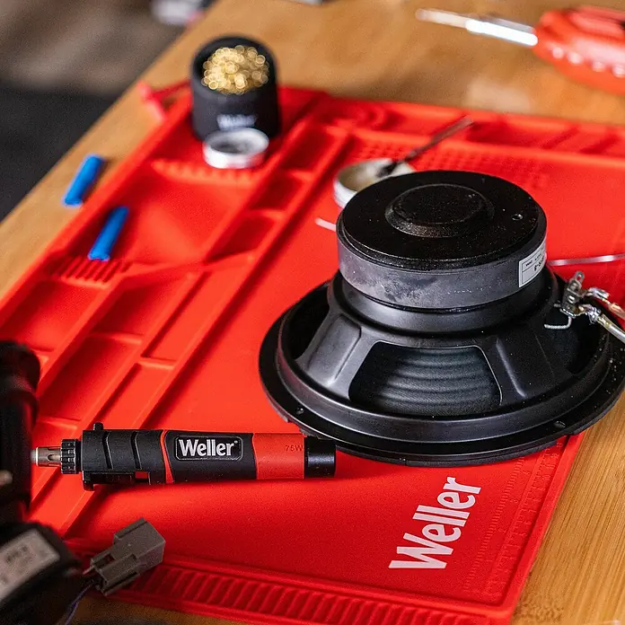 Weller Gaslötkolben-Set WLBUK75 - 9 -tlg., Arbeitstemperatur: 454 °C - Ein Weller Lötkolben und ein Lautsprecher liegen auf einer roten Weller-Matte. Perfekt für Reparaturen und Bastelarbeiten.