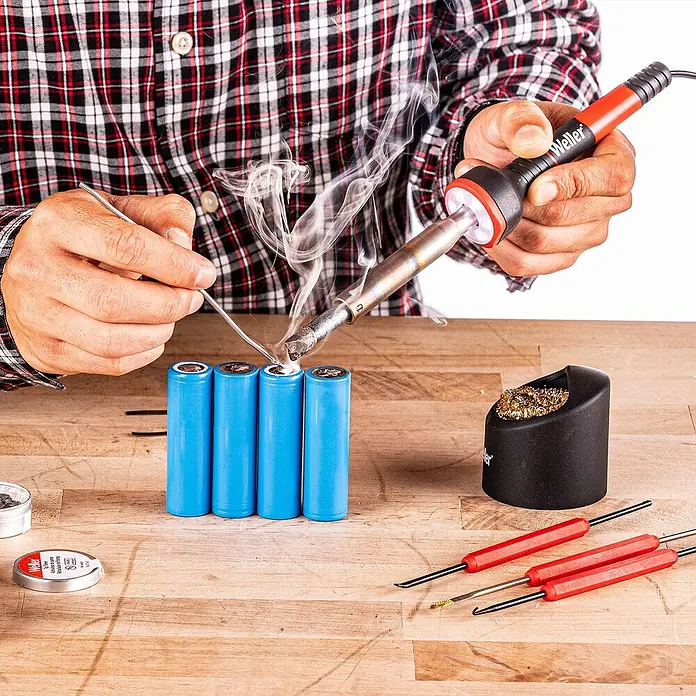 Weller Lötkolben-Set WLIRK6023C - 6 -tlg., Arbeitstemperatur: 427 °C, 60 W - Ein Mann lötet mit einem Weller-Lötkolben eine Batterie. Rauch steigt auf. Werkzeuge liegen auf dem Tisch.