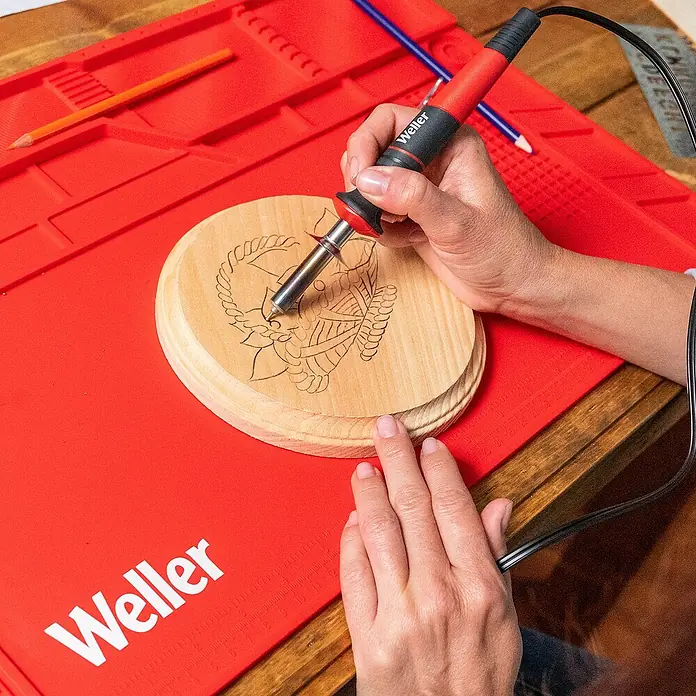 Weller Brandmal-Set WLIWBK2523C - Gesamtumfang: 15 -tlg. - Die Weller Lötstation wird zum Gravieren eines Designs auf ein Holzstück verwendet, das auf einer roten Silikonmatte liegt.