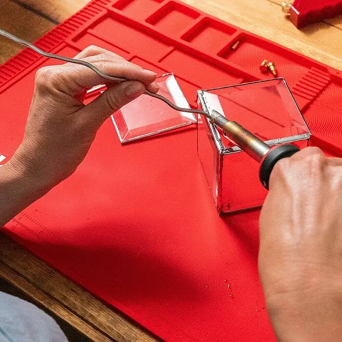 Weller Lötkolben-Set WLIRPK8023C - 6 -tlg., Arbeitstemperatur: 485 °C - Nahaufnahme eines Handwerkers, der Glas mit einem Lötkolben auf einer roten Unterlage lötet, um eine transparente Box zu erstellen.
