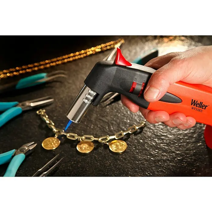 Weller Lötbrenner WT13EU - Arbeitstemperatur: 1.300 °C - Nahaufnahme einer Weller WT13EU-Lötlampe beim Erhitzen einer goldenen Halskette mit Münzanhängern, umgeben von Werkzeugen auf dunkler Oberfläche.