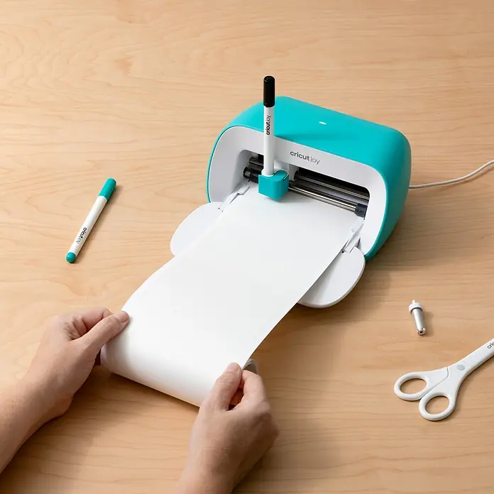 Cricut Joy Aufkleber Smart Label Auflösbar1 Stk., Weiß, 13,9 x 90 cm - Nahaufnahme eines Cricut Joy Schneideplotters mit Stiften, Zubehör und einer Person, die eine weiße Vinylrolle handhabt.