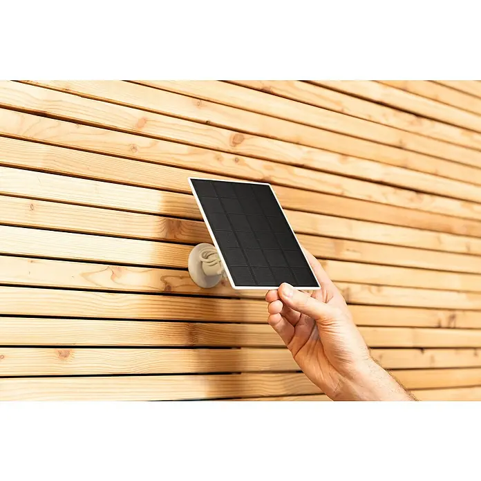 Abus Solarmodul PPIC91600 W PRO - L x B x H: 12 x 1 x 17 cm, IP65 - Nahaufnahme eines montierten Solarpanels mit einer Hand, die es vor einer Holzwand hält, Hervorhebung seiner Installation und Funktionalität.