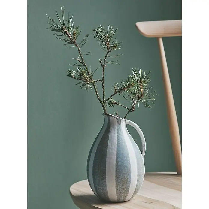 Alpina Wandfarbe Feine Farben - Held des Waldes, Matt, 2,5 l - Gestreifte Krugvase aus Keramik mit einem Kiefernzweig darauf, die eine natürliche und ruhige Atmosphäre schafft. Dekorative und funktionelle Vase.