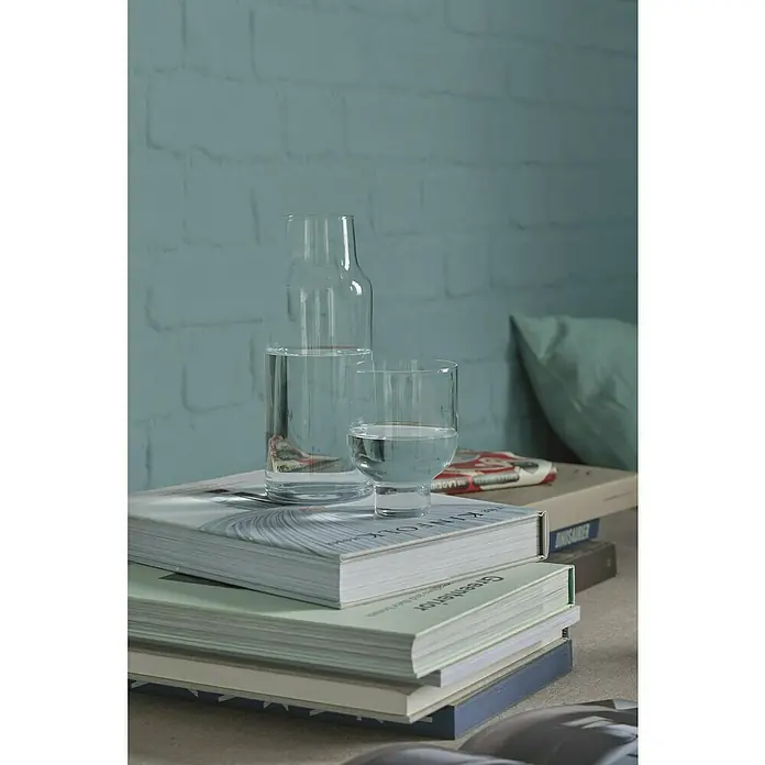 Alpina Wandfarbe Feine Farben - Quelle der Gelehrten, Matt, 2,5 l, Konservierungsmittelfrei - Klares Glas mit Wasser steht auf einem Stapel Bücher vor einer strukturierten, hellblau gestrichenen Wand. Das Glas und die Bücher harmonieren gut.