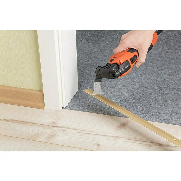 Wolfcraft Tauchsägeblatt Expert BiM - Arbeitsbreite: 50 mm - Oszillierendes Elektrowerkzeug schneidet eine goldene Bodenschiene vor einer Tür. Das Werkzeug ist orange und schwarz.