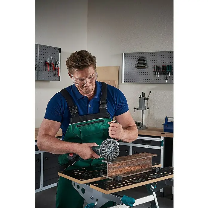 Wolfcraft Scheiben-Drahtbürste - Stahldraht, Durchmesser: 115 mm, Gezopft - Ein Mann benutzt eine Drahtbürste an einem Werkstück auf einer Werkbank. Er trägt eine Schutzbrille und Arbeitskleidung.