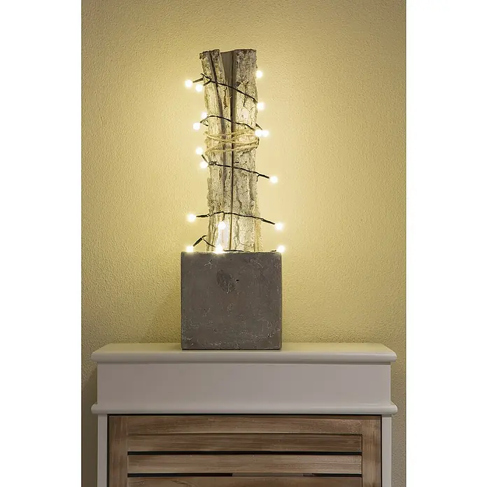 Konstsmide LED-Lichterkette - Innen, Lichtfarbe: Warmweiß, Anzahl Leuchtmittel: 50 Stk., Batteriebetrieben - Nahaufnahme einer dekorativen Baumstammlampe auf einem weißen Sideboard, die eine gemütliche Atmosphäre schafft.