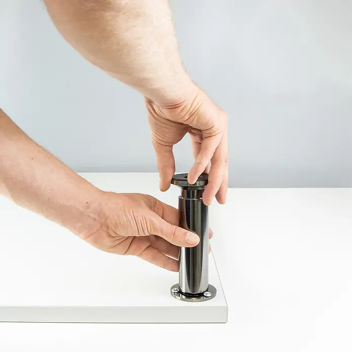 Wagner System Möbelfuß - L x B x H: 60 x 60 x 115 mm, Ø x H: 3 cm x 115 mm, Aluminium, Höhenverstellung: 10 cm - 11,5 cm - Ein silberfarbenes, höhenverstellbares Tischbein wird von zwei Händen auf einer weißen Oberfläche montiert.