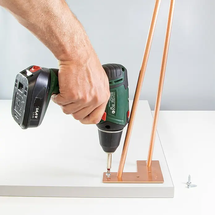 Wagner System Möbelfuß Hairpinlegs - Stahl, Höhe: 20 cm, Gold - Ein Akku-Bohrschrauber befestigt eine roségoldene Tischbeinhalterung auf einer weißen Tischplatte. Schrauben liegen bereit.