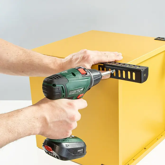 Wagner System Möbelfuß - Höhe: 12 cm, Stahl, Schwarz - Ein Akku-Bohrschrauber befestigt eine schwarze Halterung an einem gelben Metallkasten. Präzision und Kraft vereint für optimale Ergebnisse.