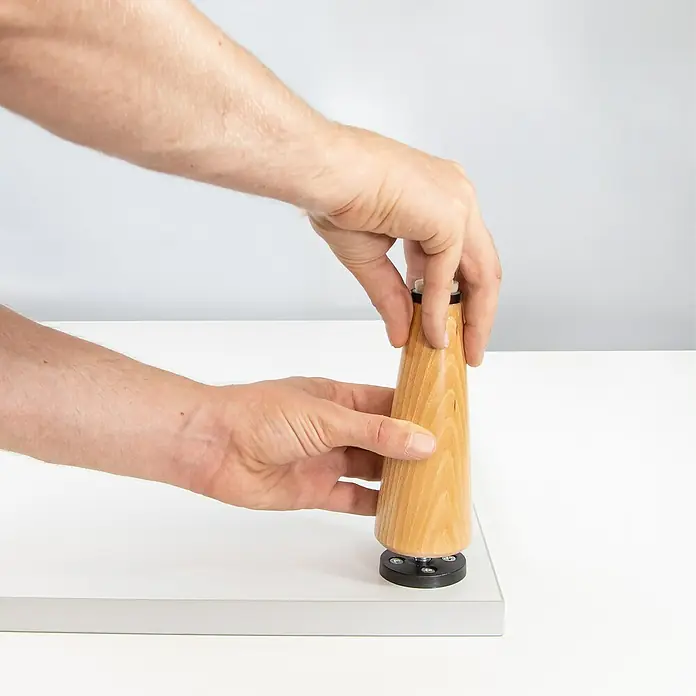 Wagner System Möbelfuß - Ø x H: 75 x 55 mm, Holz - Eine Hand befestigt ein konisches Holzbein an einer weißen Oberfläche mit einer schwarzen Halterung, wodurch das Produkt hervorgehoben wird.