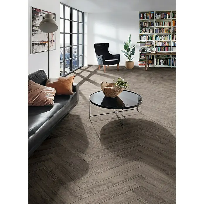 Krono Original Laminat Herringbone - Rutherford Oak, Fischgrät, 630 x 126 x 8 mm - Wohnzimmer mit dunklem Holzboden im Fischgrätmuster, schwarzem Ledersofa, Sessel und Bücherregal. Der Boden verleiht dem Raum eine elegante Note.