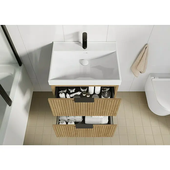 Camargue Badmöbel-Set Yasumi60 cm, Hamilton Eiche - Aufsicht auf einen Waschtischunterschrank mit zwei offenen Schubladen, gefüllt mit Badutensilien, in einem modern eingerichteten Badezimmer.