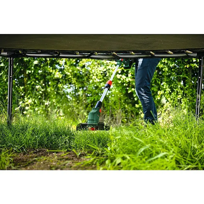 Bosch Power for All 18V Akku-Rasentrimmer UniversalGrassCut 18V-26-50018 V, Li-Ionen, Ohne Akku - Eine Person trimmt Gras unter einem Trampolin mit einem grünen und silbernen Bosch Akku-Rasentrimmer, der mit einer roten Akzentfarbe versehen ist.