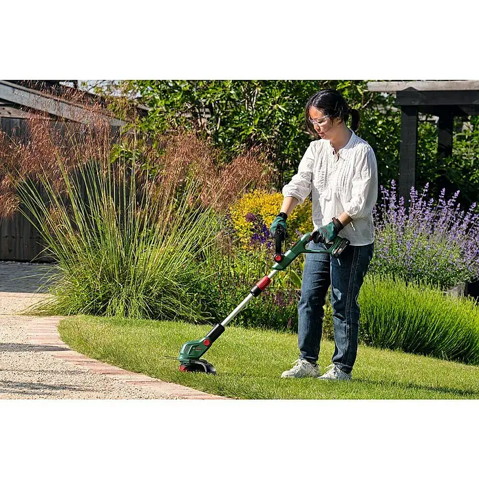 Bosch Power for All 18V Akku-Rasentrimmer UniversalGrassCut 18V-26-50018 V, Li-Ionen, Ohne Akku - Frau schneidet Rasen mit einer Bosch Akku-Grasschere. Die Grasschere ist grün, schwarz und rot und hat einen Akku.