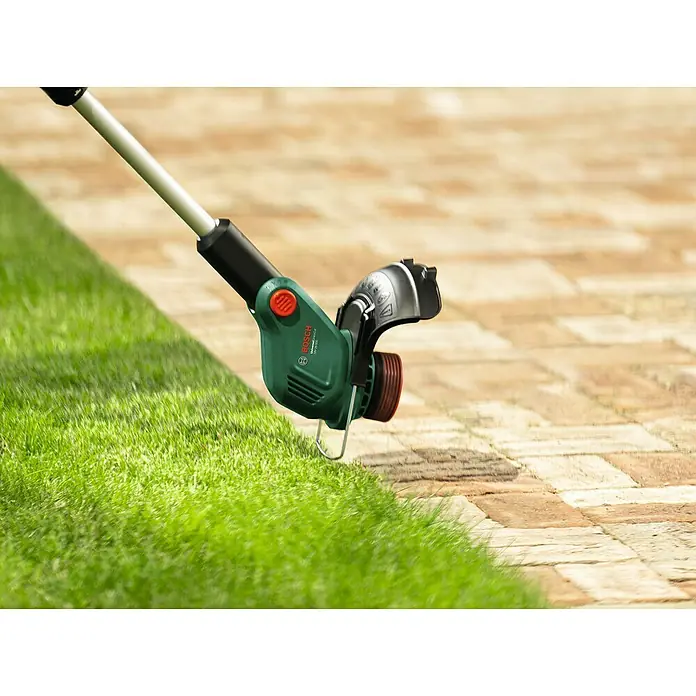 Bosch Power for All 18V Akku-Rasentrimmer UniversalGrassCut 18V-23-450 - 18 V, Ohne Akku - Akku-Rasentrimmer von Bosch schneidet Gras entlang eines Bürgersteigs. Das Gerät ist grün und hat einen roten Knopf.