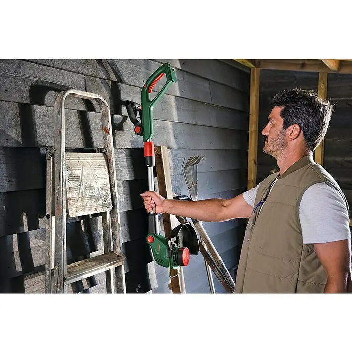Bosch Power for All 18V Akku-Rasentrimmer UniversalGrassCut 18V-23-450 - 18 V, Ohne Akku - Ein Mann hält einen grünen Rasentrimmer von Bosch vor einer hölzernen Wand, neben einer Leiter und Gartengeräten.