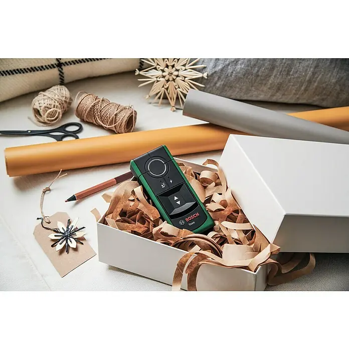 Bosch Detector Truvo II - Apto para: Detectar cables eléctricos y metal, Profundidad de detección: Máx. 70 mm de metales ferrosos - La imagen muestra un medidor de distancia Bosch Truvo en una caja de regalo, rodeado de papel de regalo, hilo y otros elementos decorativos.