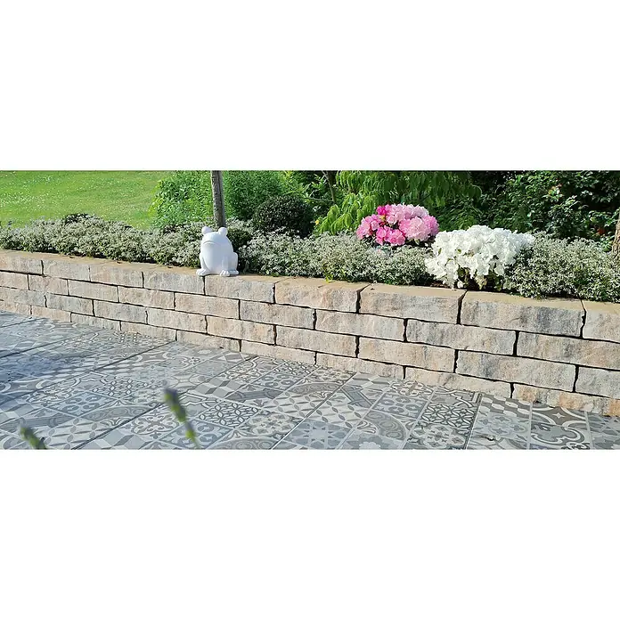 Diephaus Mauerstein Victorville - L x B x H: 40 x 20 x 20 cm, Muschelkalk, Beton - Eine niedrige Steinmauer mit einer Keramikfroschstatue und Blumen, die einen charmanten Garteneffekt erzeugen, ergänzt durch dekorative Bodenfliesen.