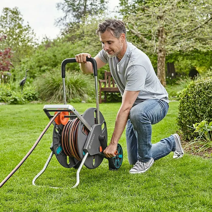 Gardena Schlauchwagen CleverRoll L Easy - Kunststoff/Metall, Schlauchkapazität: 100 m, 13 mm (½″) - Ein Mann benutzt eine Gardena Schlauchbox mit einer orangefarbenen Schlauchrolle auf einem grünen Rasen.